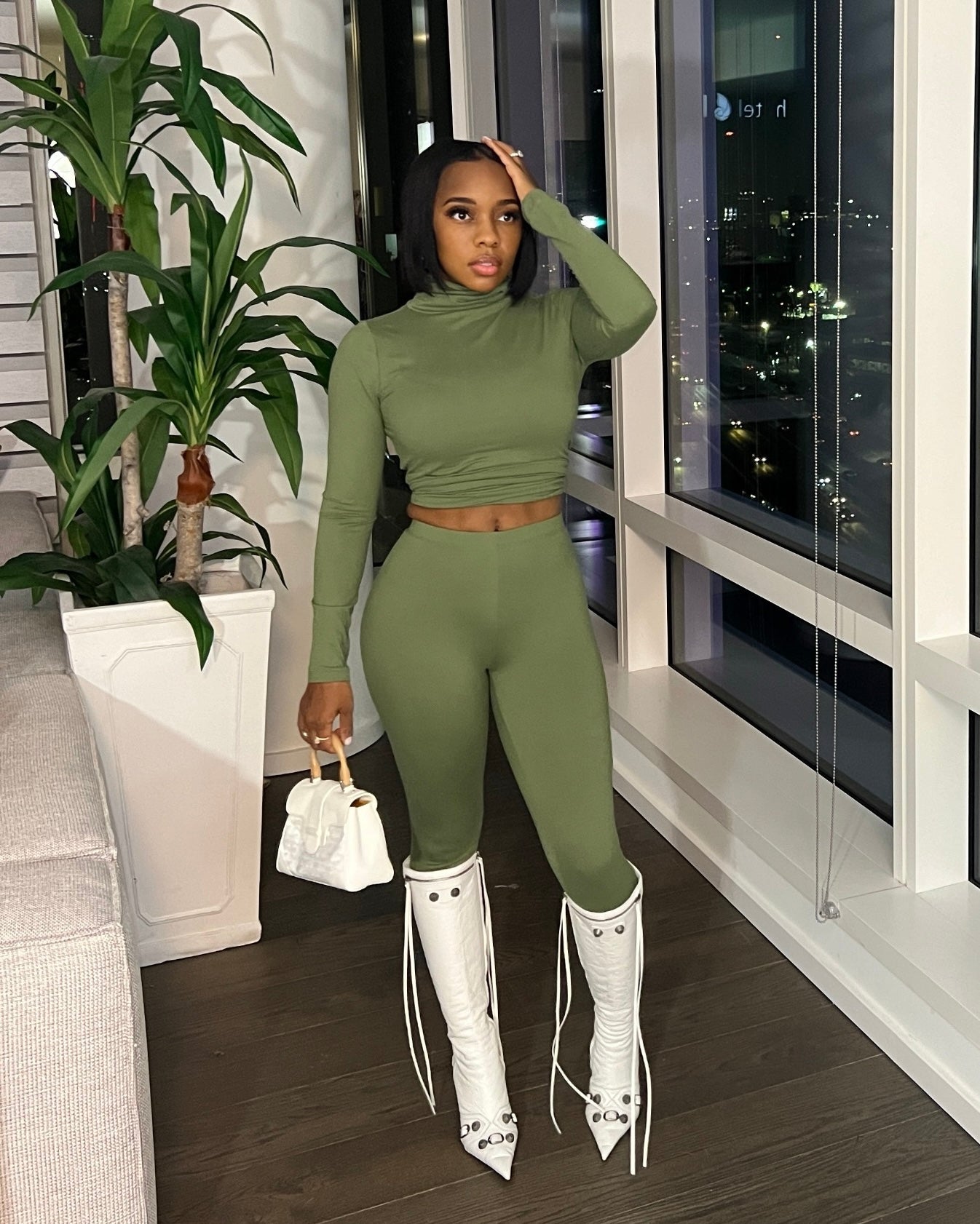 A woman wearing a green turtleneck top and matching leggings, standing in a room with a clear view of the cityscape.